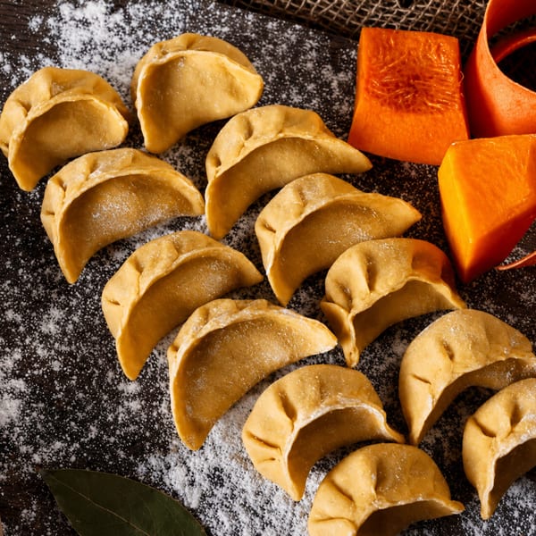 Uncooked pumpkin dumplings dusted with flour, alongside fresh pumpkin pieces on a dark surface.