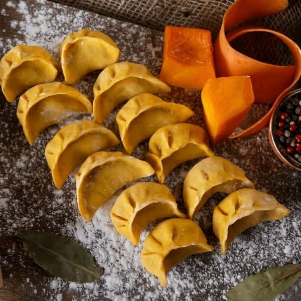 Uncooked yellow dumplings dusted with flour, alongside pumpkin pieces, a peel, and spices on a dark surface.