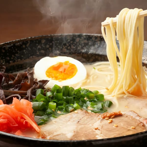 Steaming bowl of ramen with noodles lifted by chopsticks, egg, pork, scallions, and pickled ginger.